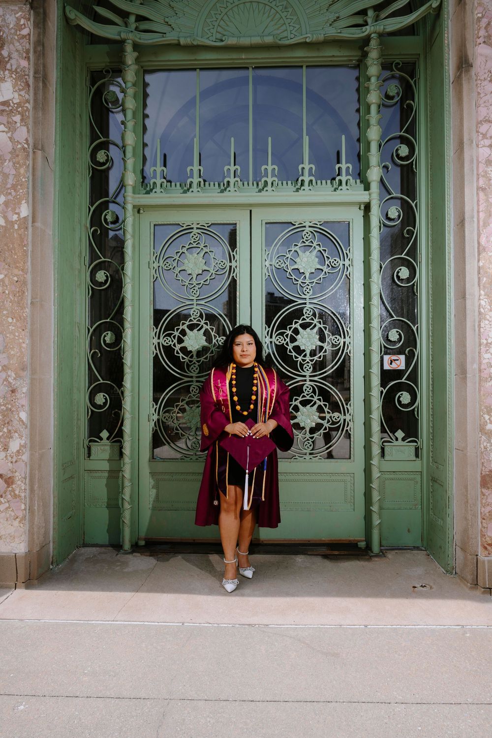 Graduate in burgundy graduation regalia poses in front of ornate green doors with decorative ironwork.