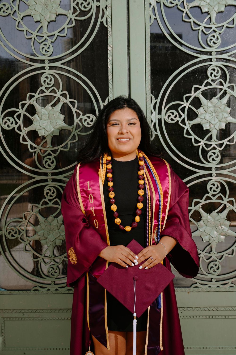Graduate in burgundy graduation regalia poses in front of ornate door wearing decorative stole and necklace.