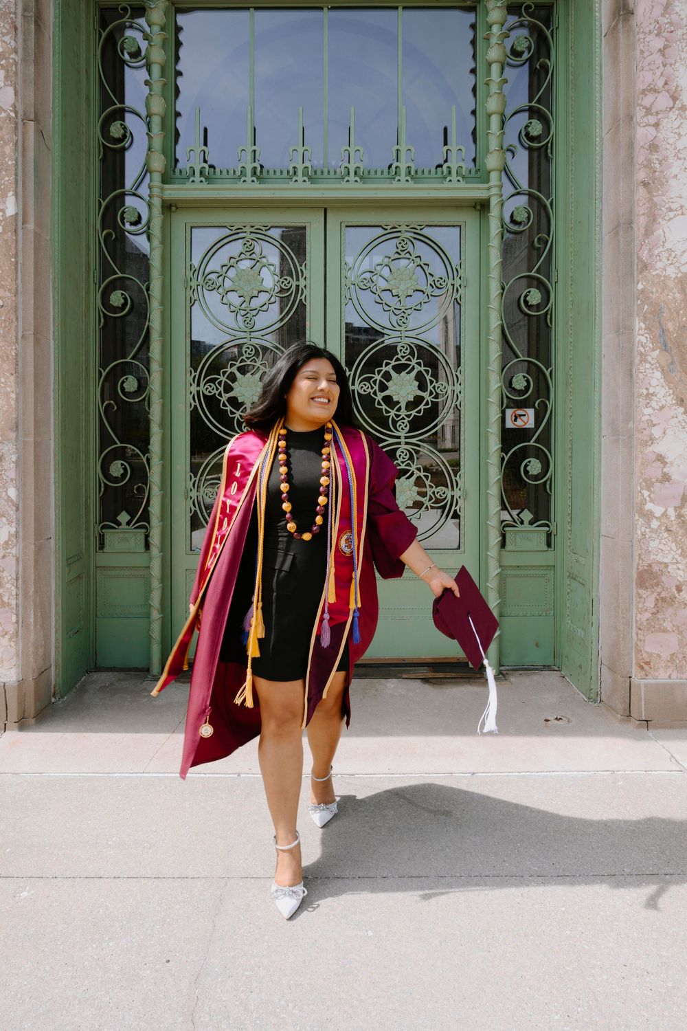 Graduate in crimson regalia and dress strikes a pose in front of vintage art nouveau doorway.