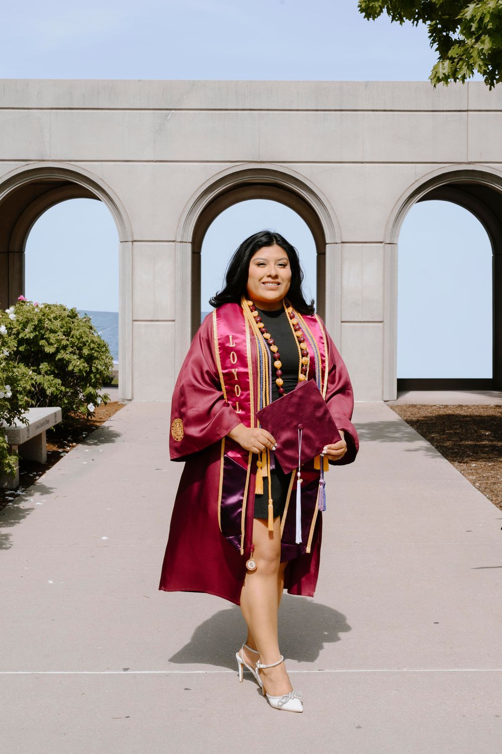 Graduate poses confidently under stone archways wearing burgundy graduation regalia near the waterfront.