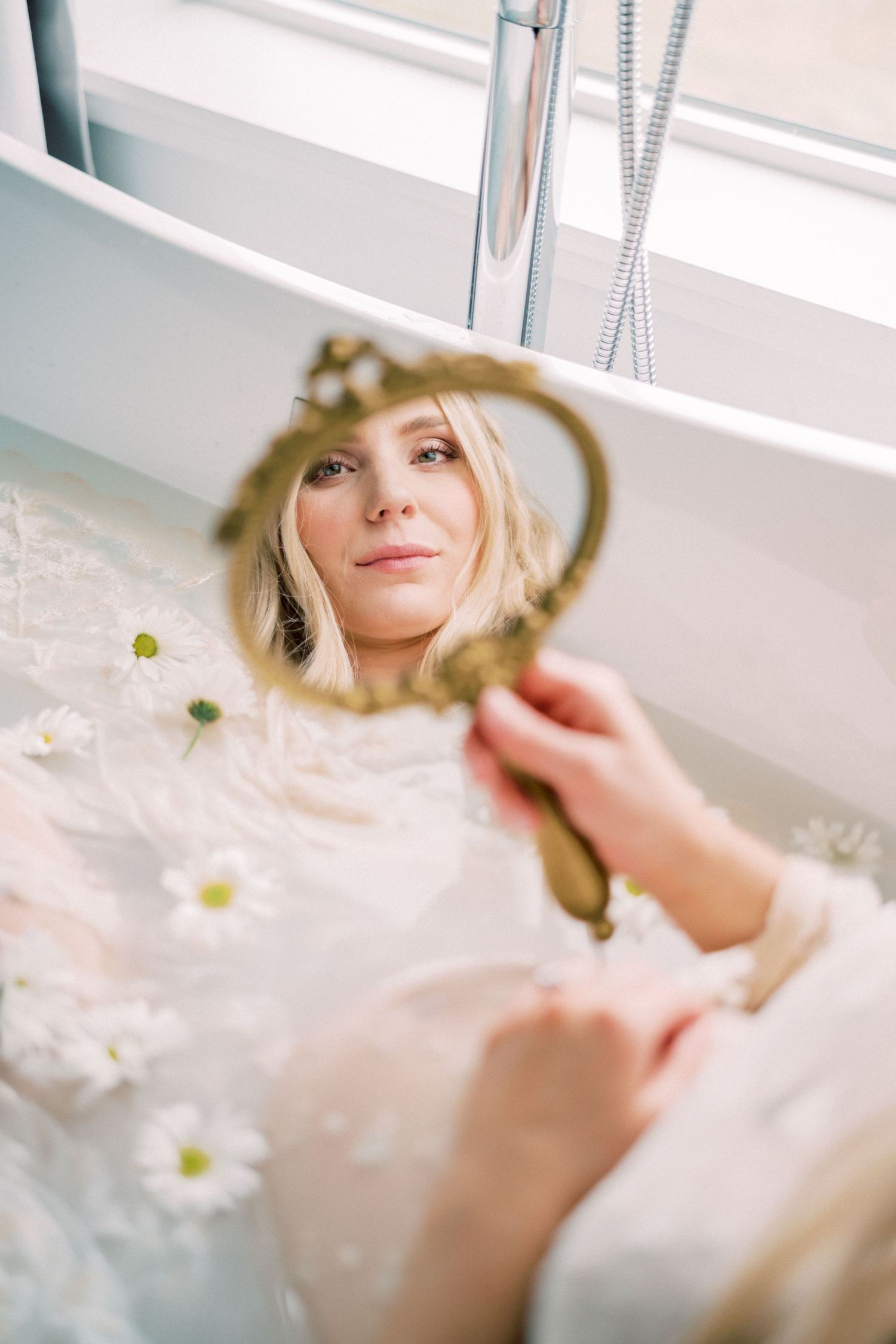 Hand holding vintage ornate gold hand mirror surrounded by delicate white daisies and soft fabrics in ethereal setting.