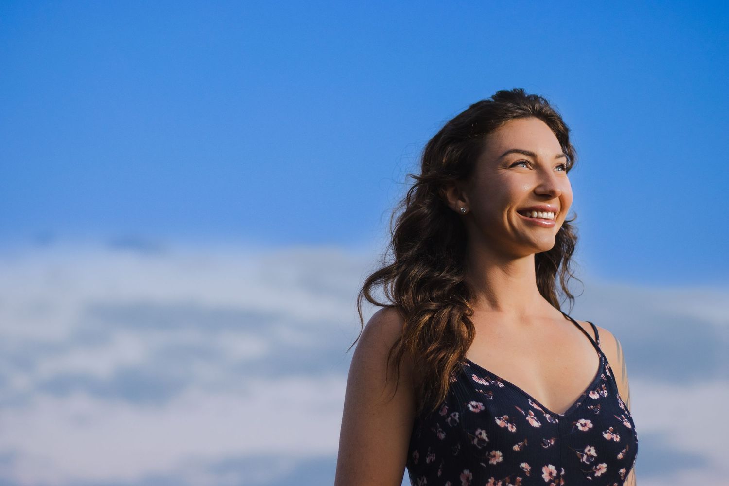 Smiling person with flowing dark hair in floral dress poses against bright blue sky with white clouds.
