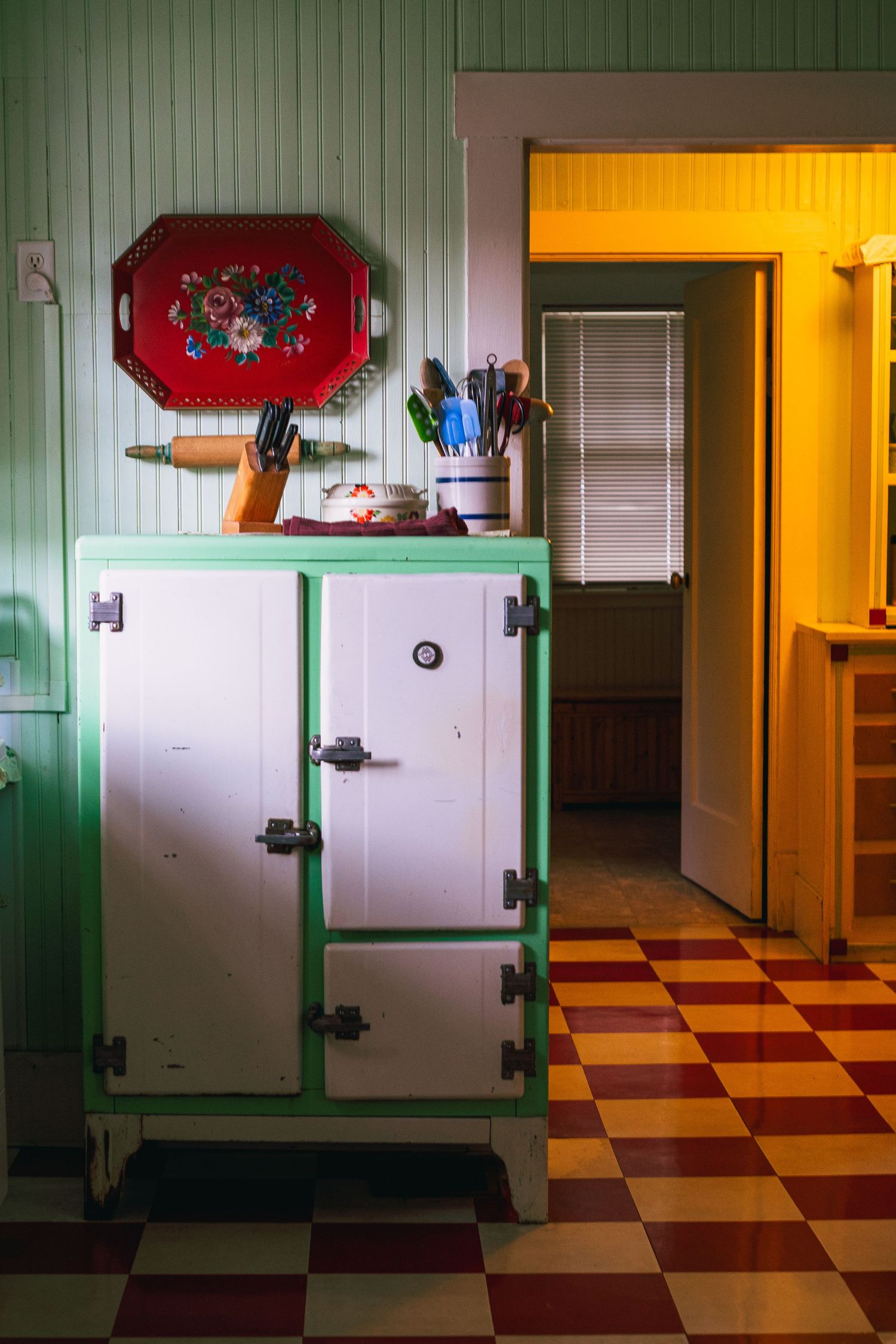 Vintage white refrigerator with green trim stands in kitchen with checkered floor and yellow doorway.