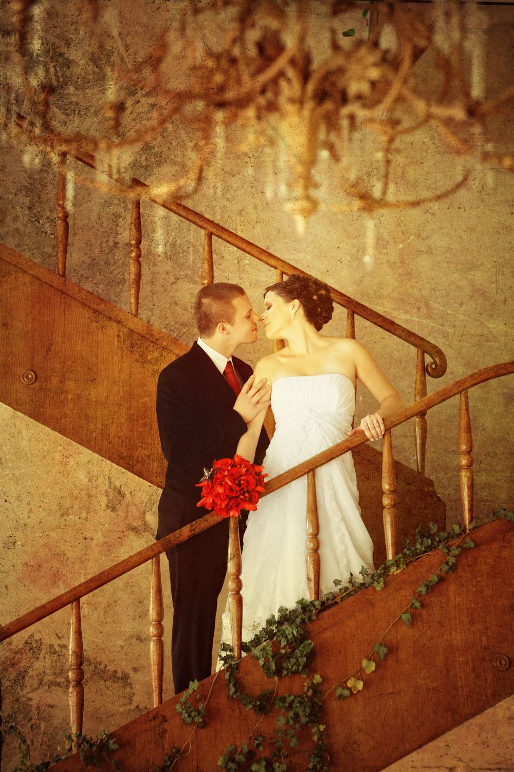 A romantic vintage wedding photo on a staircase with a bride in white holding red flowers and embracing in sepia tones.