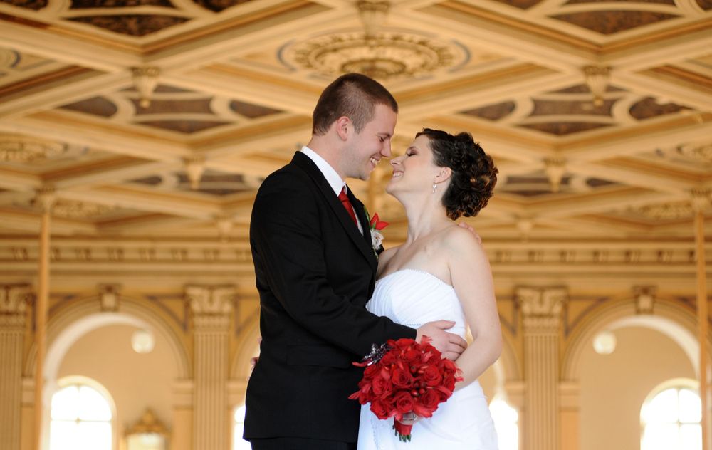 A couple shares a romantic moment in an ornate ballroom with red roses and elegant architectural details.