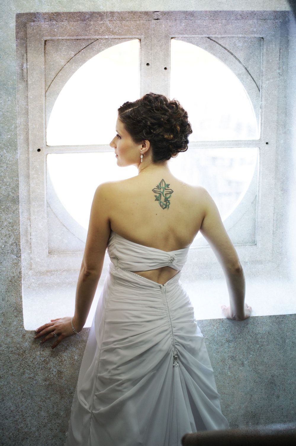 A bride in a white strapless wedding gown with a back tattoo stands by an arched window with ethereal lighting.