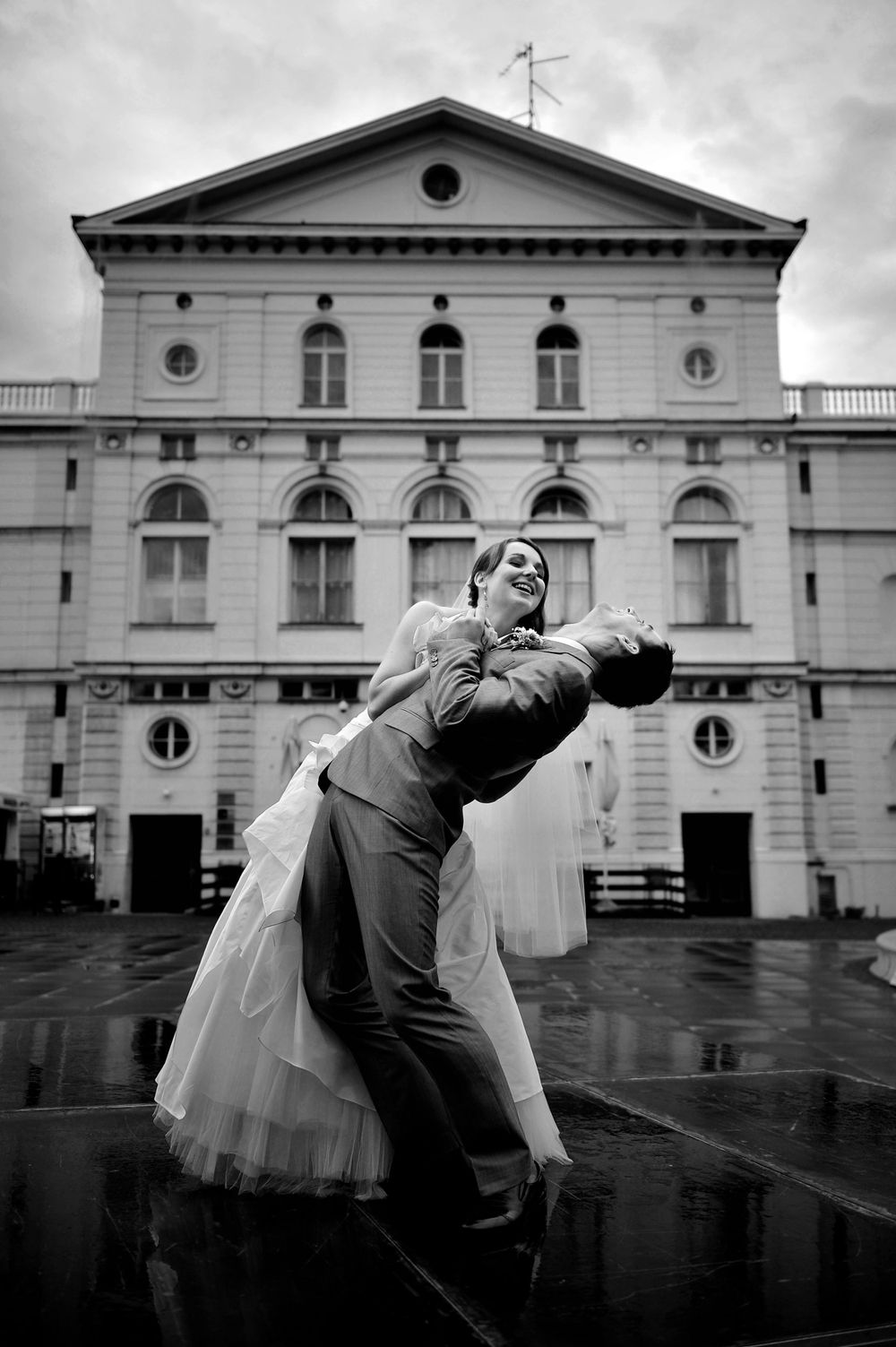 Dancers perform an elegant dip pose in front of a grand historical theater building on a rainy day in black and white.