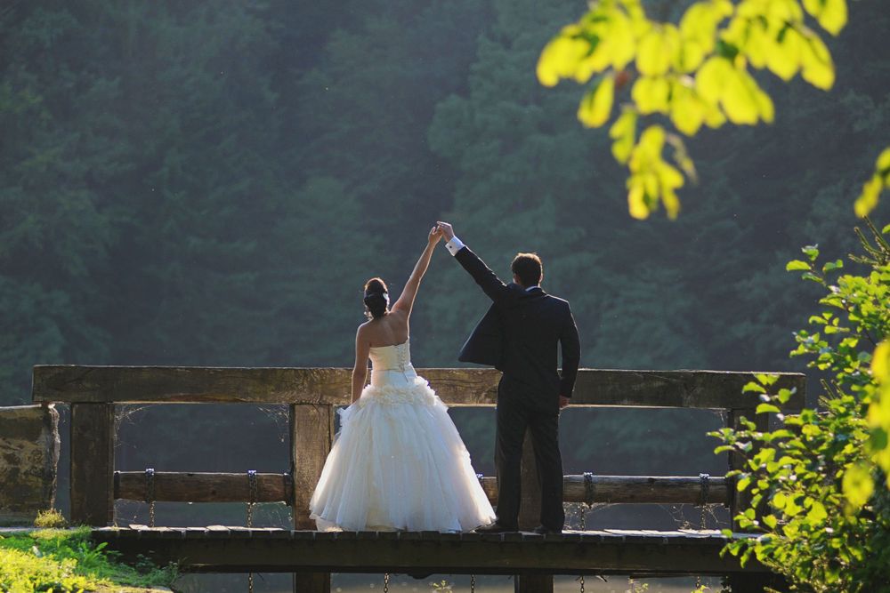 A couple in wedding attire stands on a wooden deck overlooking a scenic forest view at sunset.