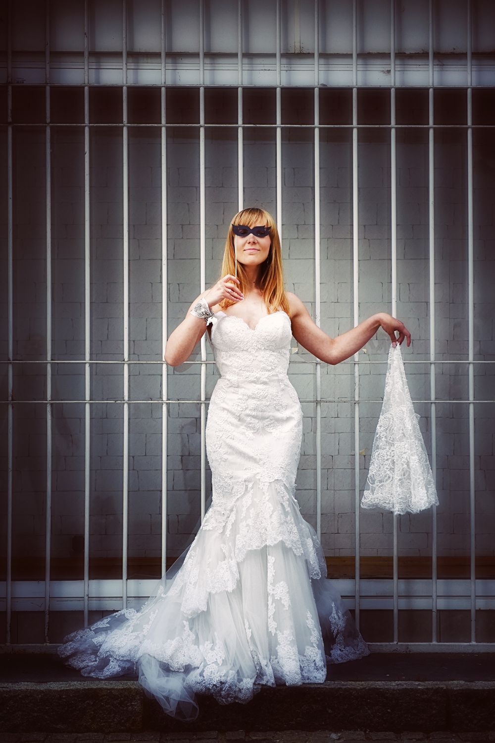 A bride in a fitted white lace mermaid wedding gown and sunglasses poses against an industrial metal backdrop.