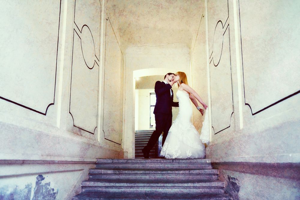 A romantic wedding couple embraces on an elegant marble staircase with ornate white walls.
