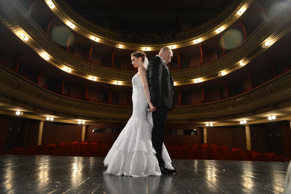 Couple in wedding attire stands on elegant theater stage with tiered balconies.