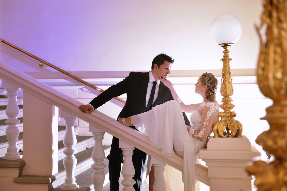 A couple in formal wedding attire share a romantic moment on a grand white staircase under elegant lighting.