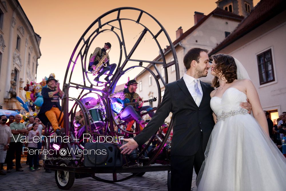 A wedding couple poses at dusk in front of a large illuminated purple spider sculpture on a European street.