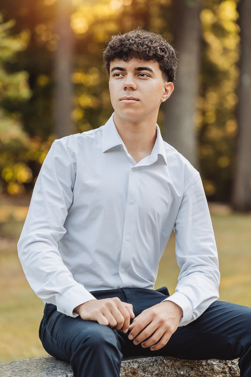 Young person in white dress shirt and dark pants sits casually on stone surface in autumn outdoor setting.