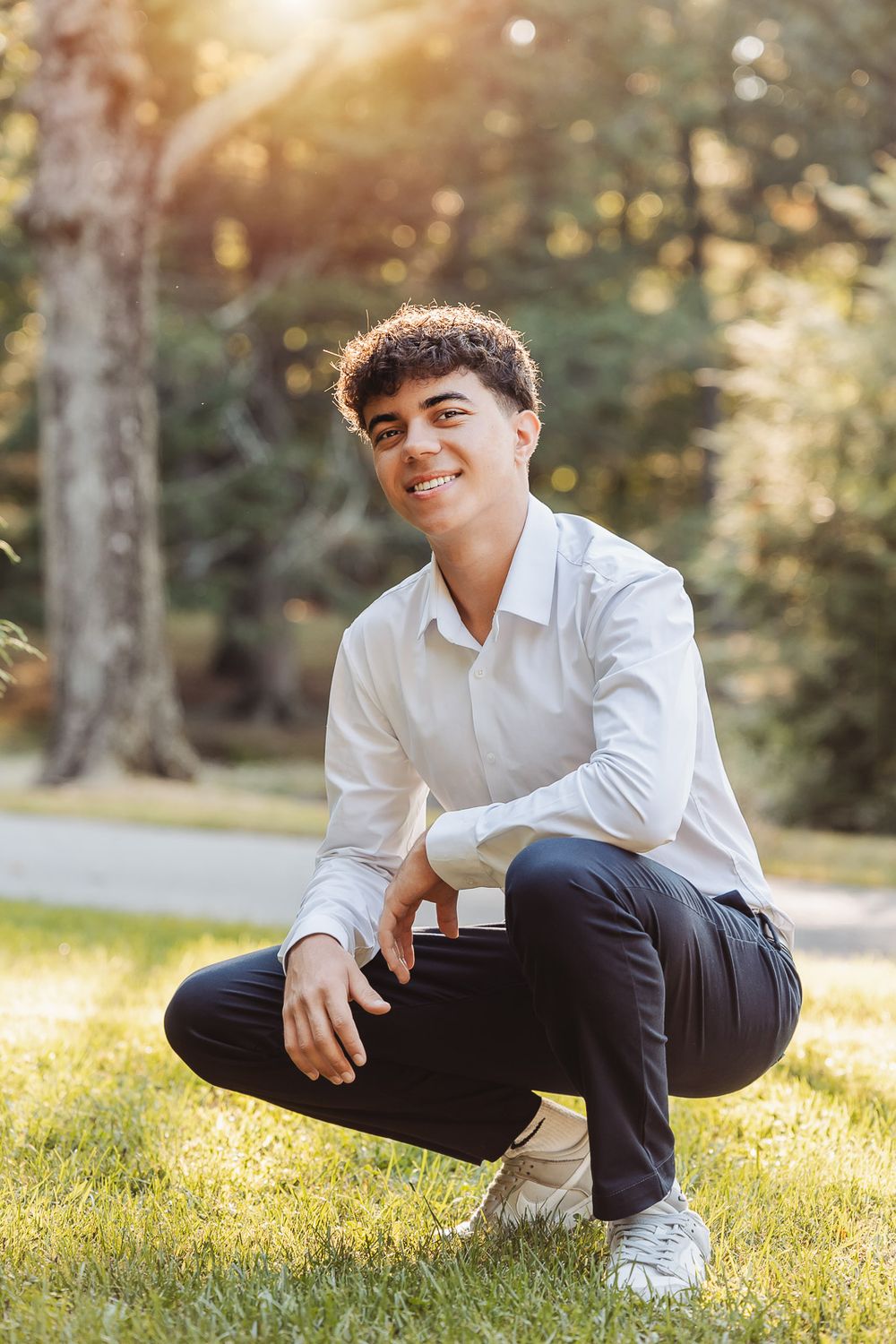 Casual portrait of someone crouching on grass in white shirt and dark pants during golden hour in a park.
