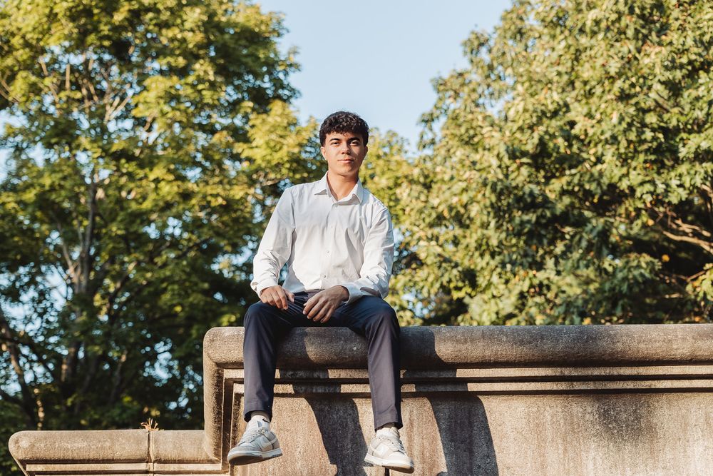 A person sits casually atop a stone wall outdoors with sunlit green trees in the background on a bright autumn day.