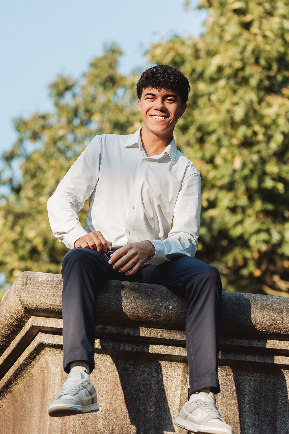 Young person in white button-down shirt sits casually on stone ledge outdoors on sunny day with trees in background.