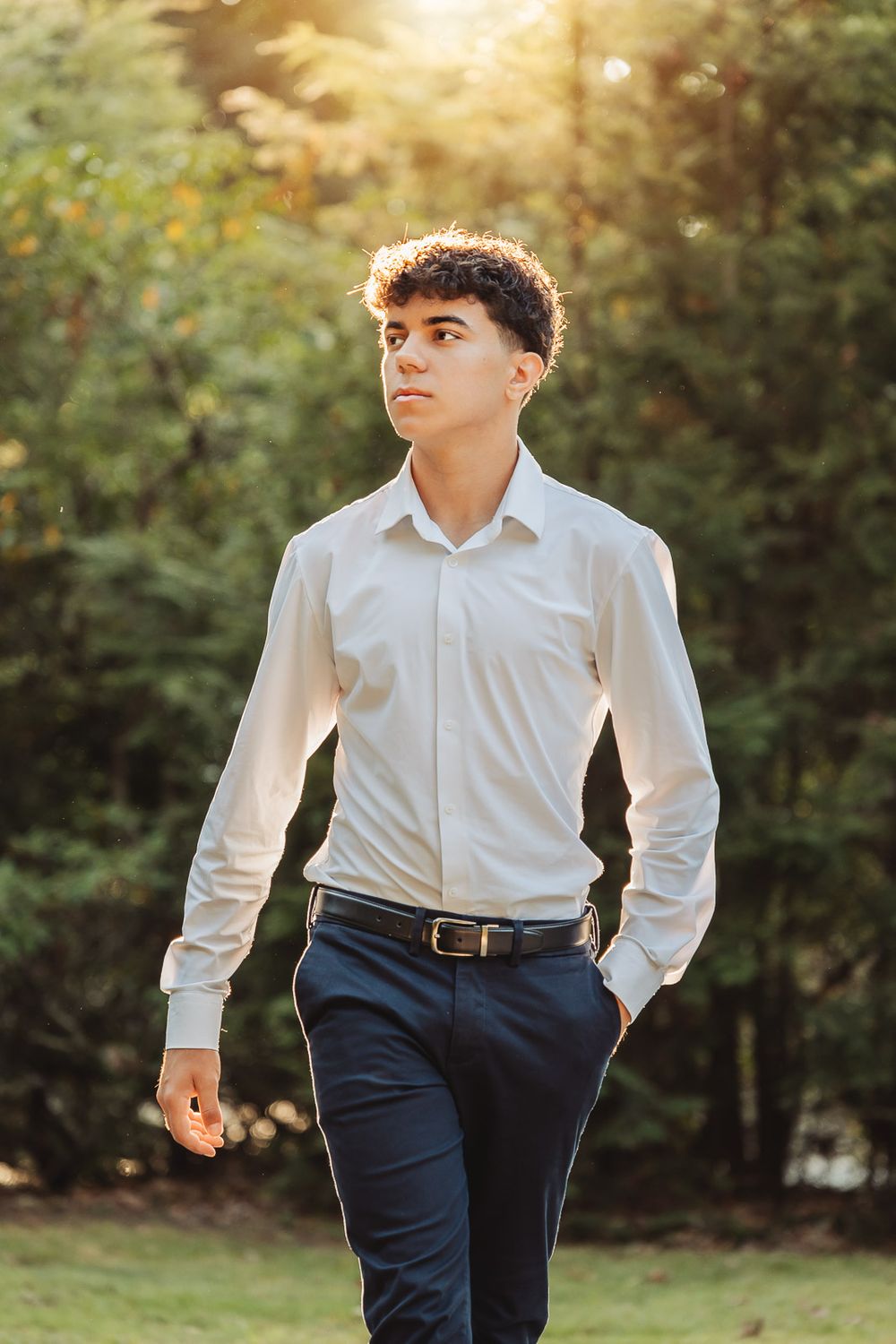 Outdoor portrait of person in white dress shirt and navy pants standing in golden sunlight with natural background.