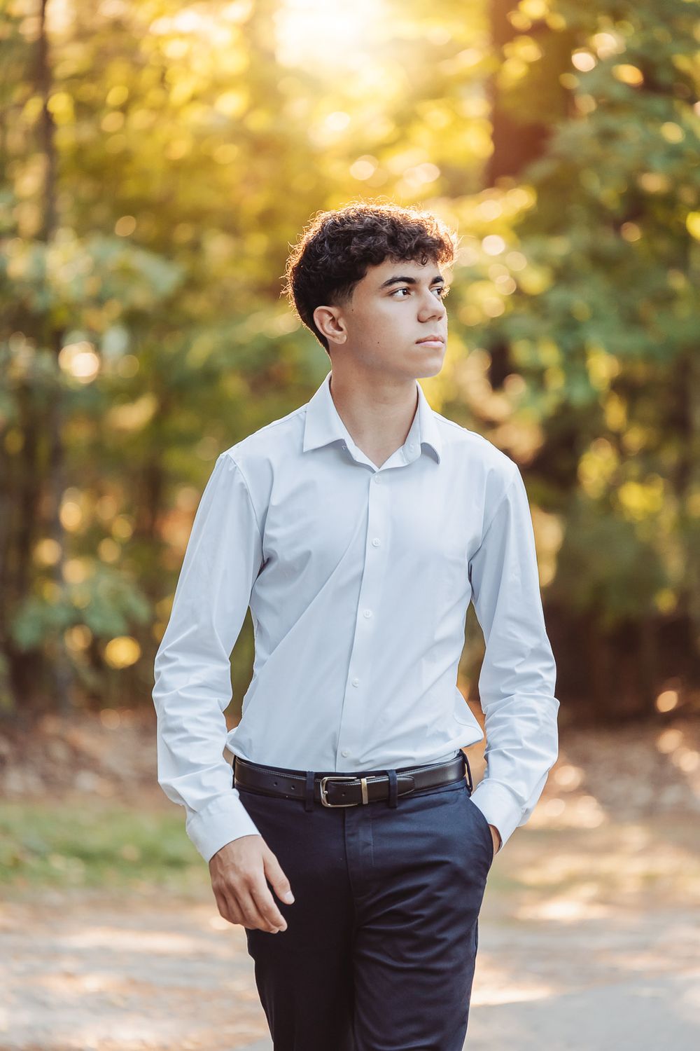 Portrait of a young person in a white dress shirt and dark pants standing outdoors with golden autumn foliage in background.