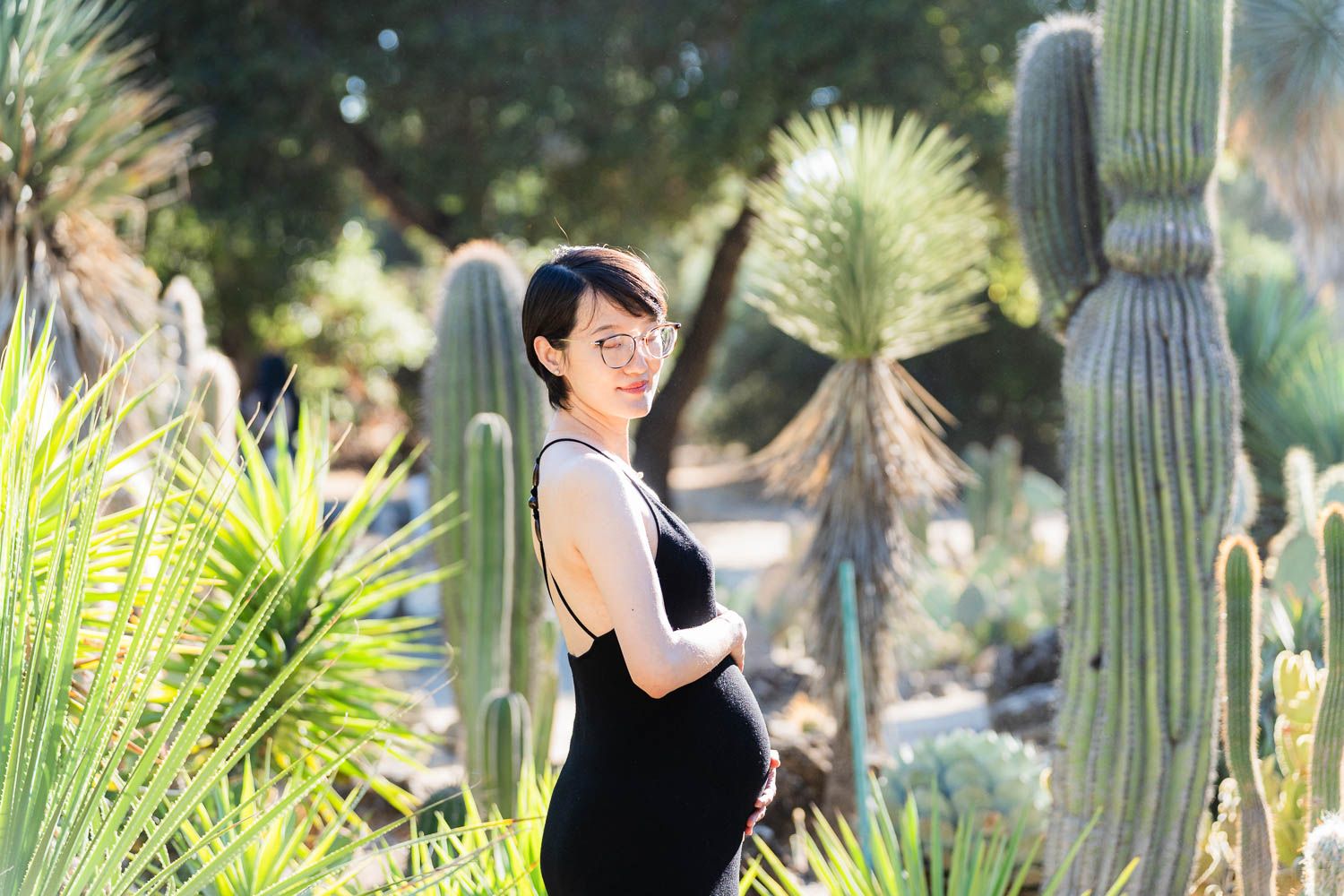 A person in black attire poses amid desert plants and tall saguaro cacti in a sunny botanical garden setting.