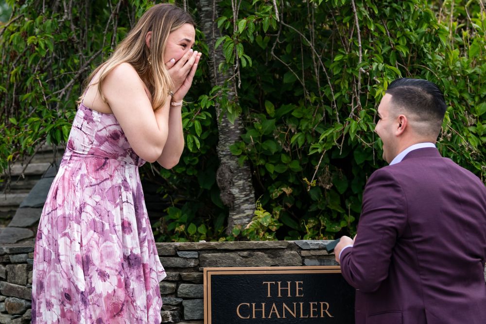 A person in a burgundy suit kneels in front of someone wearing a floral pink dress beside a sign reading The Chanler.