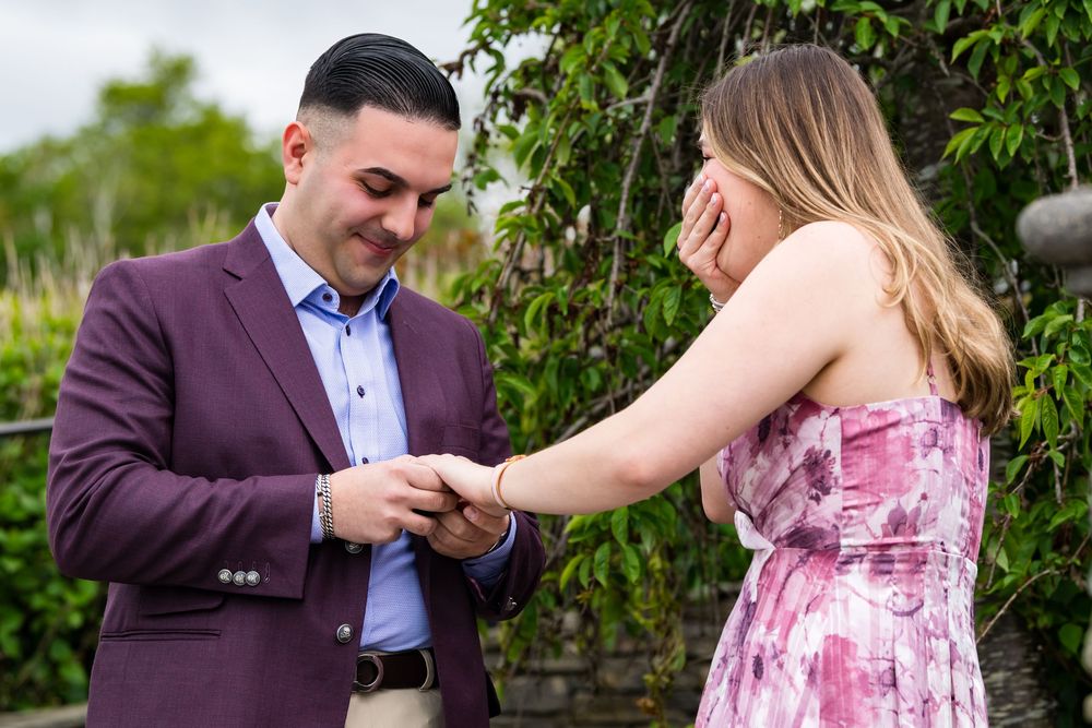 A proposal moment in an outdoor garden setting as someone in a burgundy suit presents a ring.