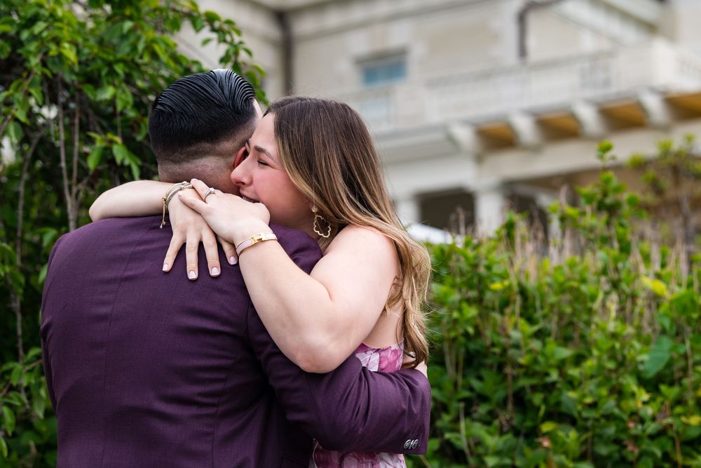 A couple in purple attire embrace intimately in a garden setting with a building visible in the background.