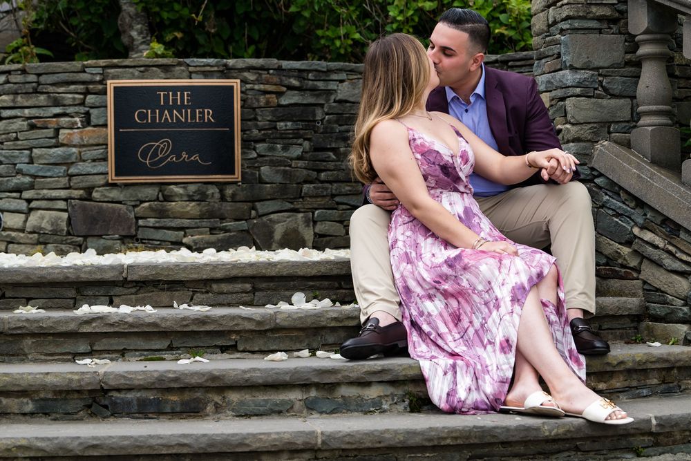 A couple sits on stone stairs near a sign for The Chamber, with the person in a floral dress leaning towards their partner.