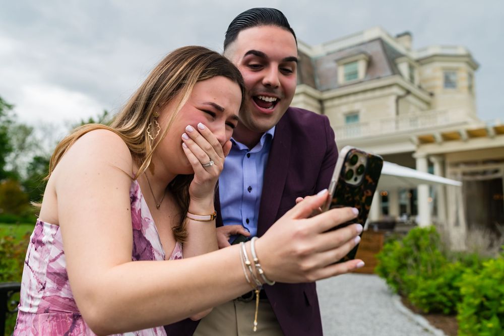A couple shares an excited moment while looking at a phone screen in front of a Victorian-style house.