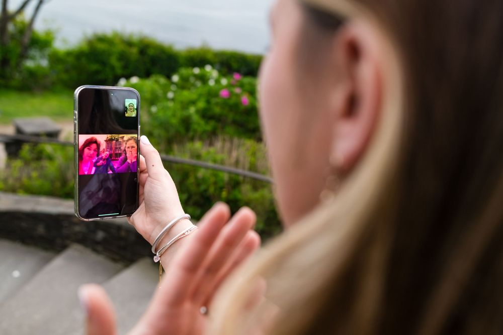 Someone using a smartphone to take a photo of purple flowers in a garden setting from over-the-shoulder perspective.