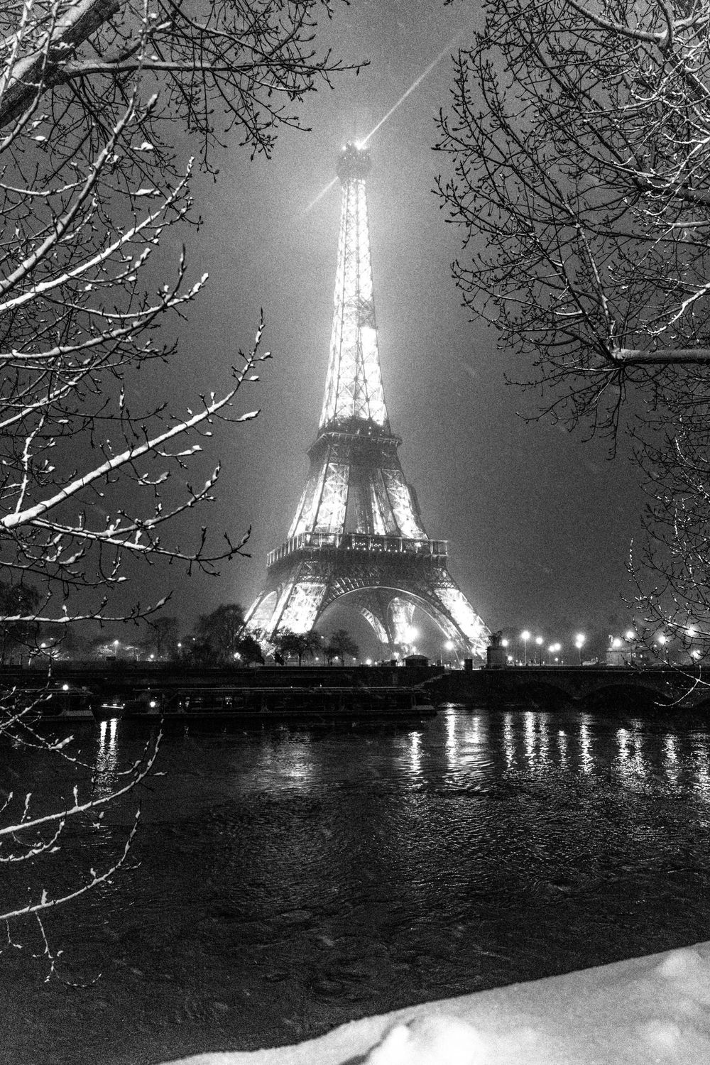 Eiffel Tower illuminated at night with snow-covered branches and Seine River reflections in black and white.