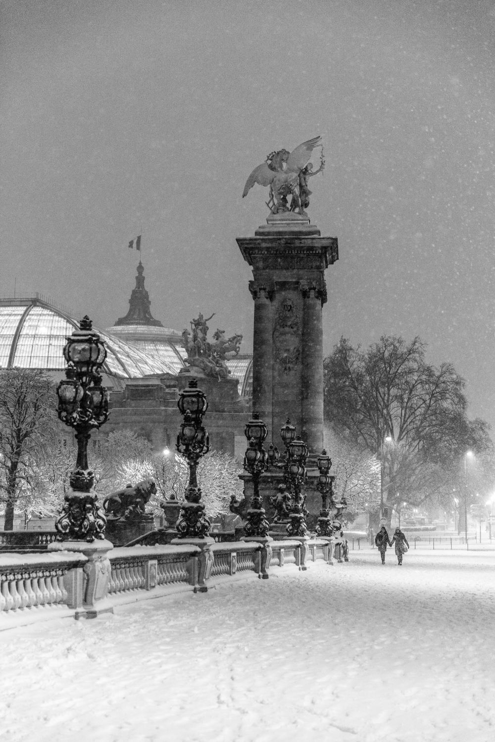 A snowy winter night scene at Pont Alexandre III in Paris with ornate lampposts and the Grand Palais visible in background.