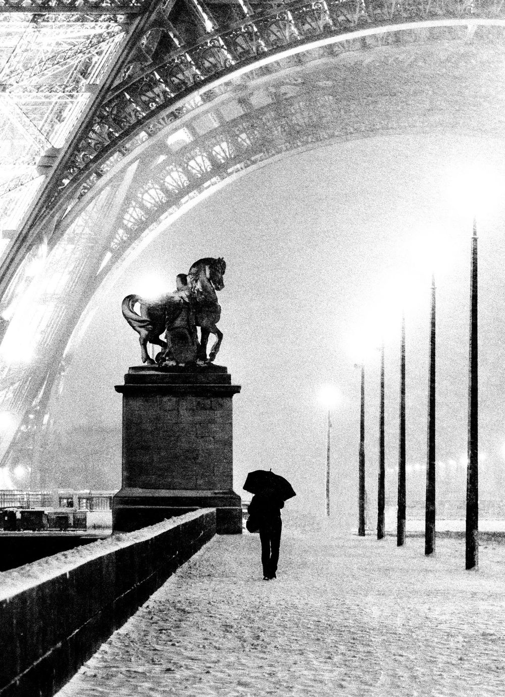 A lone figure with umbrella walks past an equestrian statue beneath the snow-covered Eiffel Tower arch.