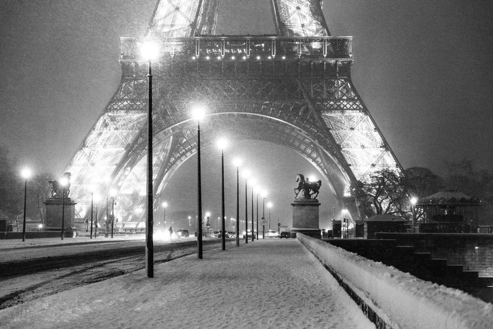 A snowy night view of the illuminated Eiffel Tower in Paris with lampposts lining a pathway beneath.