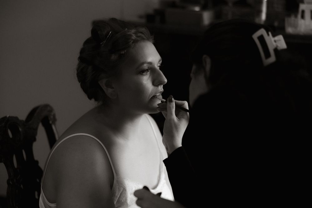 Black and white photo shows someone getting makeup applied while wearing a white tank top in dim lighting.
