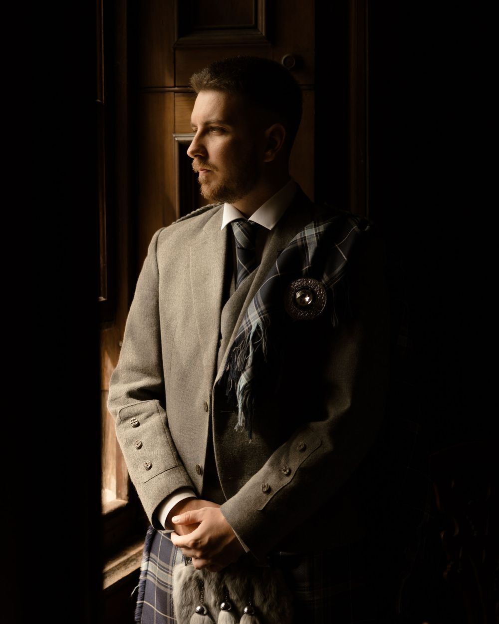 A person in a formal grey suit gazes thoughtfully through a window in dramatic lighting.