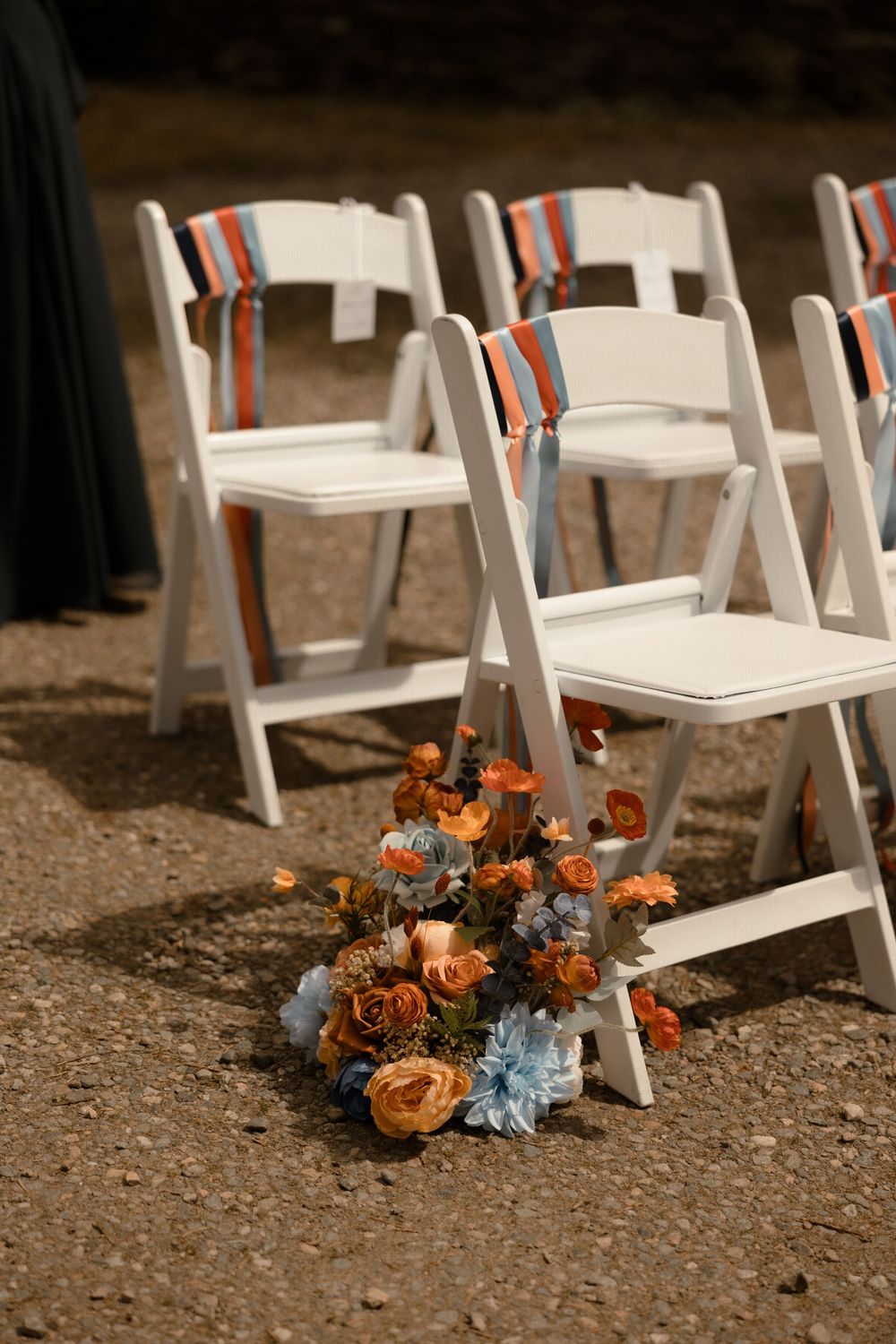 White folding chairs set up for outdoor wedding ceremony with orange and blue floral arrangements on ground.
