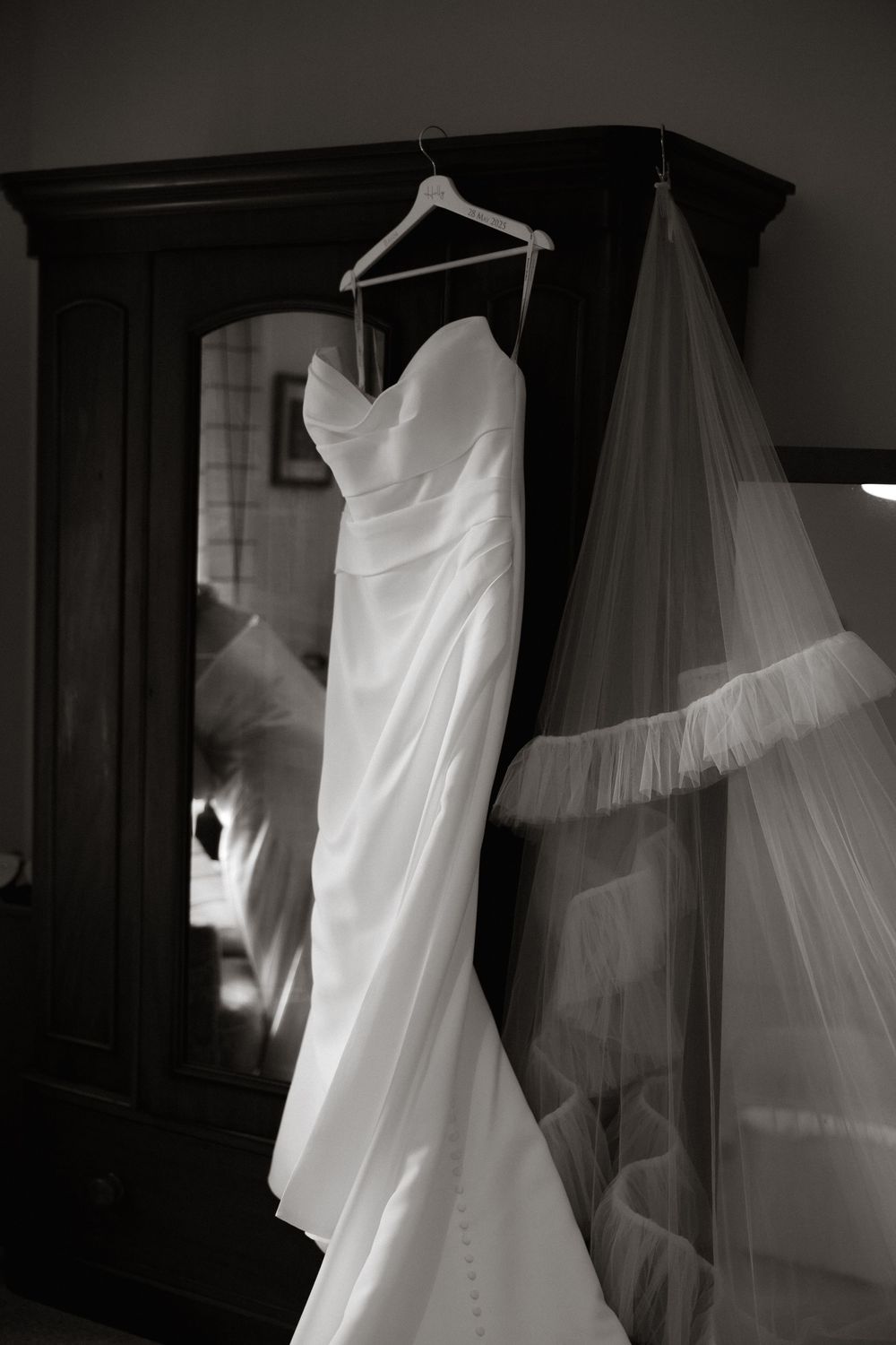 A black and white photograph of an elegant white wedding dress and veil hanging on a wardrobe.
