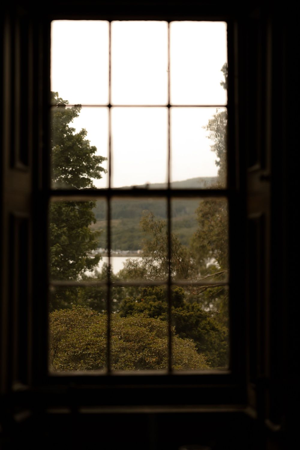 View through a dark window frame overlooking a scenic lake landscape with trees in silhouette against a bright sky.