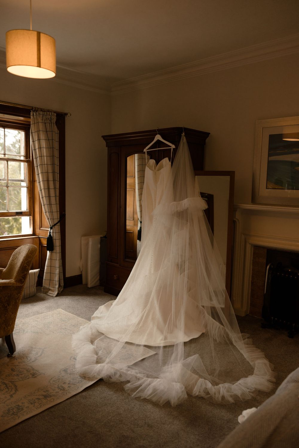 Elegant white wedding dress and flowing veil hanging from a dark wooden armoire in a sunlit bedroom with warm decor.