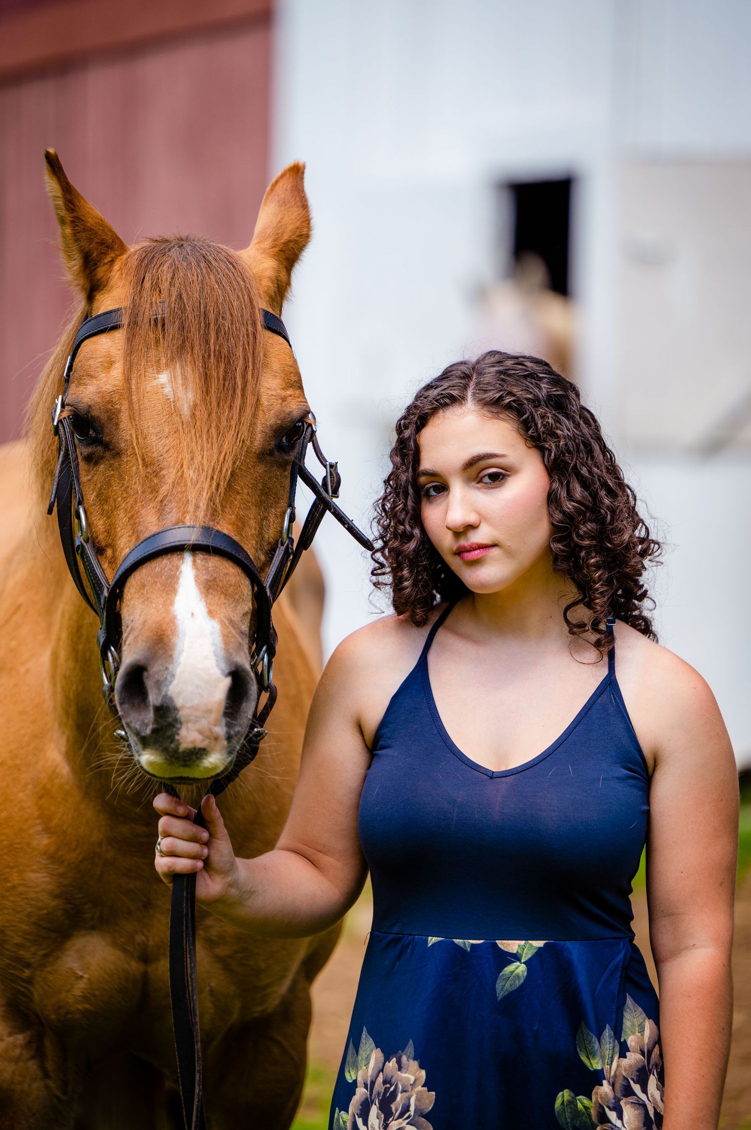 A chestnut horse wearing a bridle stands next to a young woman in a navy floral dress in front of a barn.