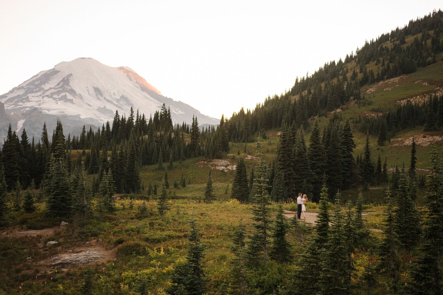 A couple embraces on a rocky outcrop overlooking a mountain valley with pine trees and golden sunset lighting.