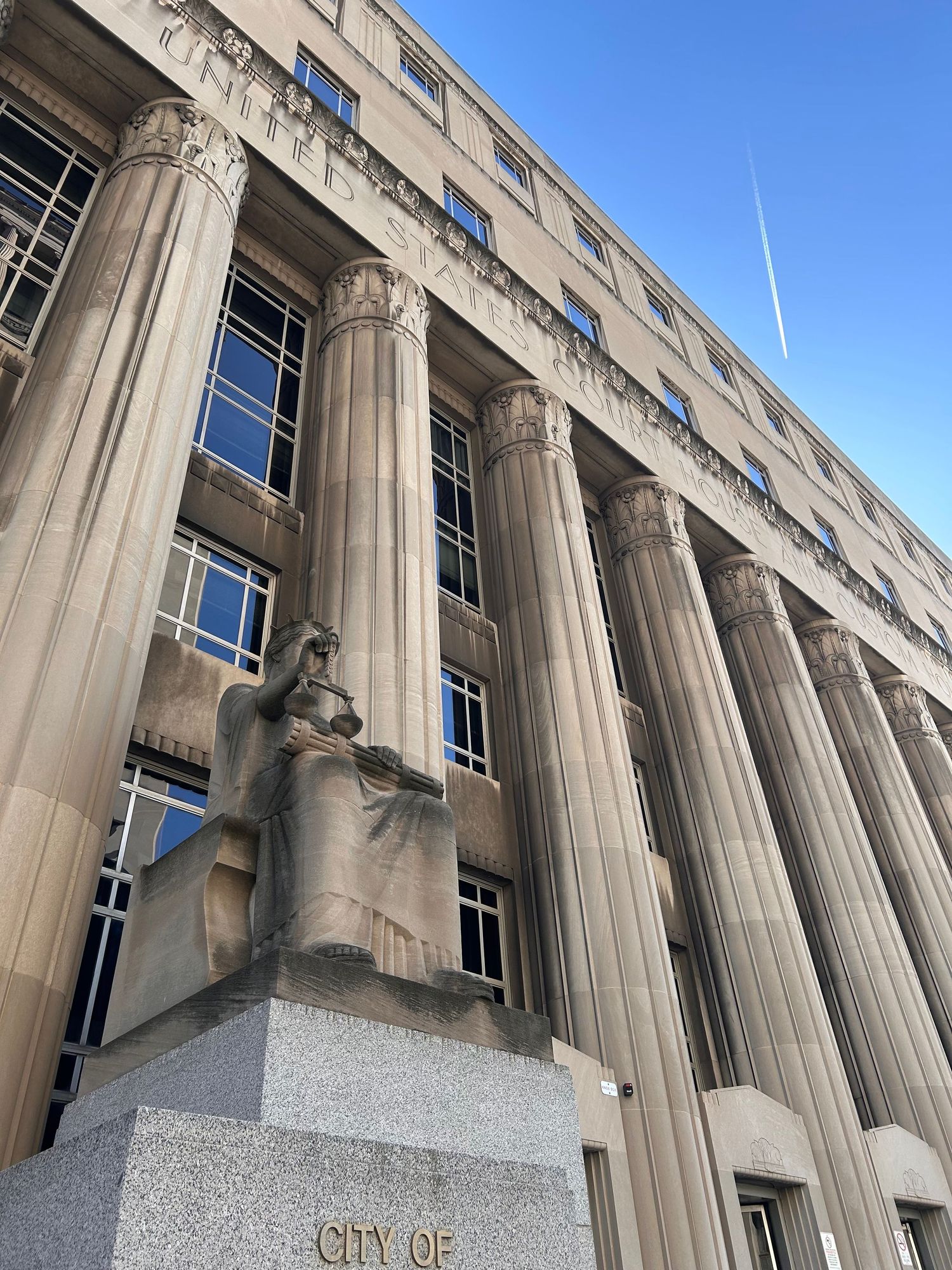 Neoclassical government building exterior with tall stone columns and a seated statue adorning its facade.
