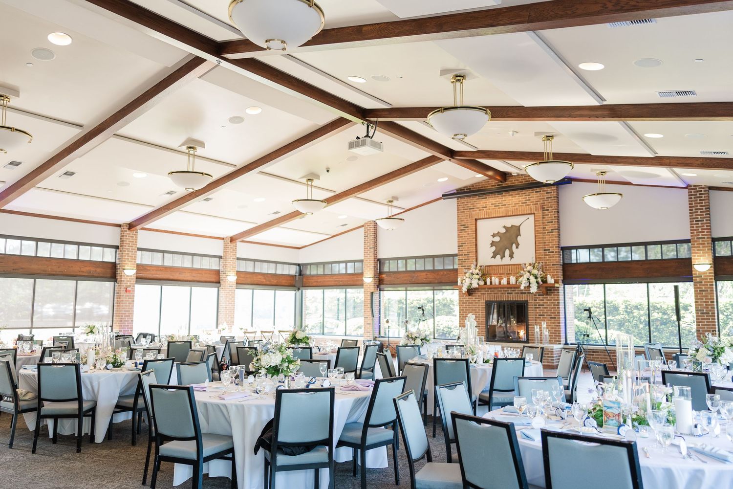 A bright wedding reception venue with exposed wooden beams, round tables, and elegant pendant lighting in a rustic-chic setting.