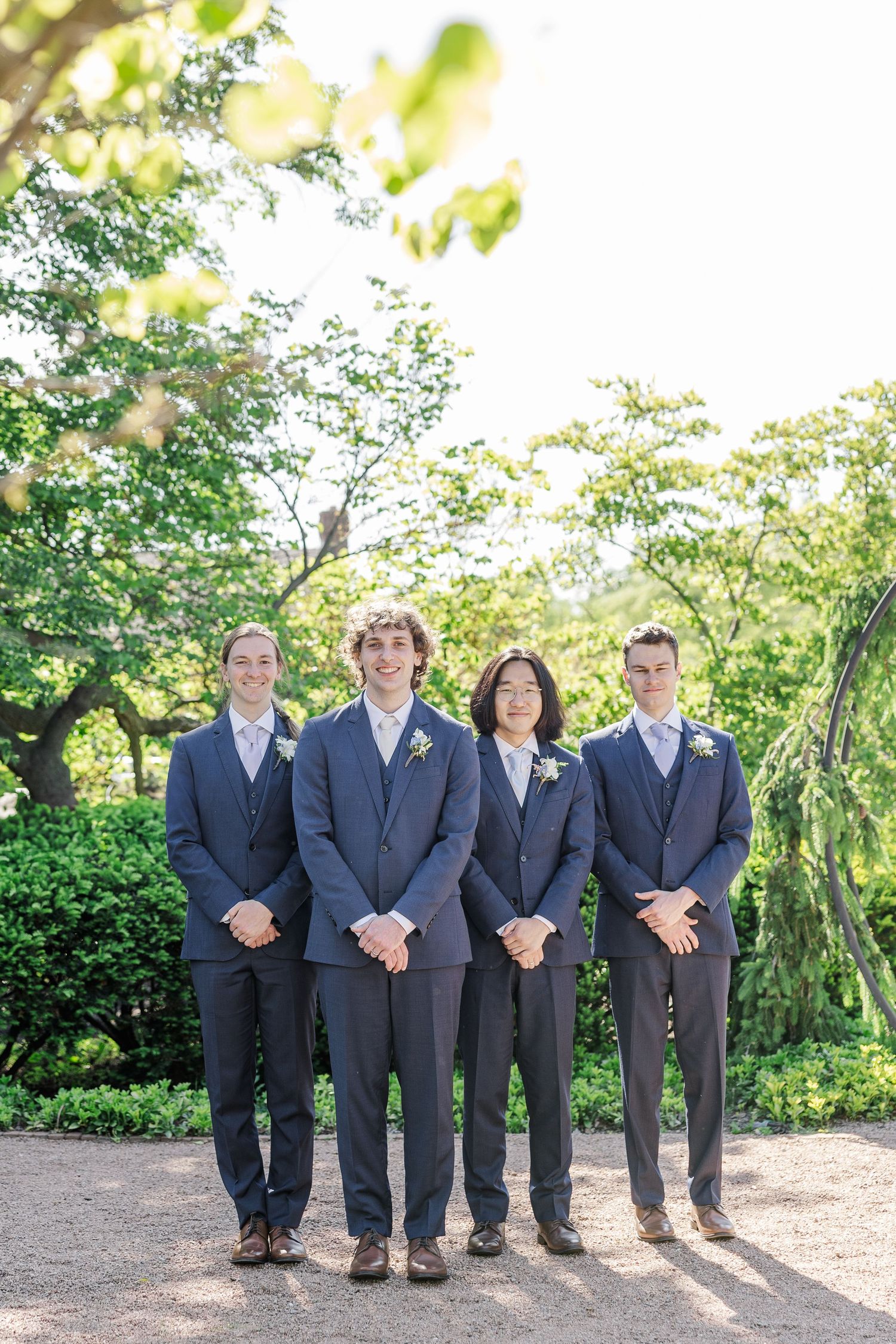 A group of four individuals wearing matching navy blue suits stand together outdoors in a garden setting.