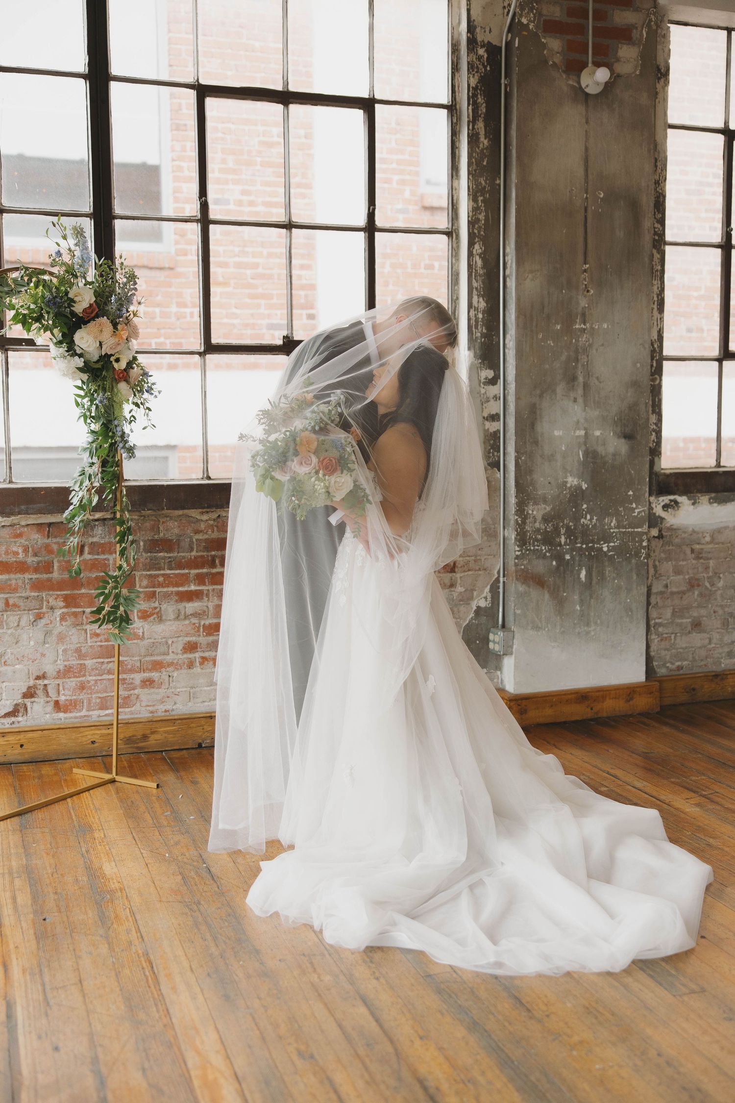 A bride in a flowing white wedding gown and veil poses in an industrial-style venue with exposed brick and hardwood floors.
