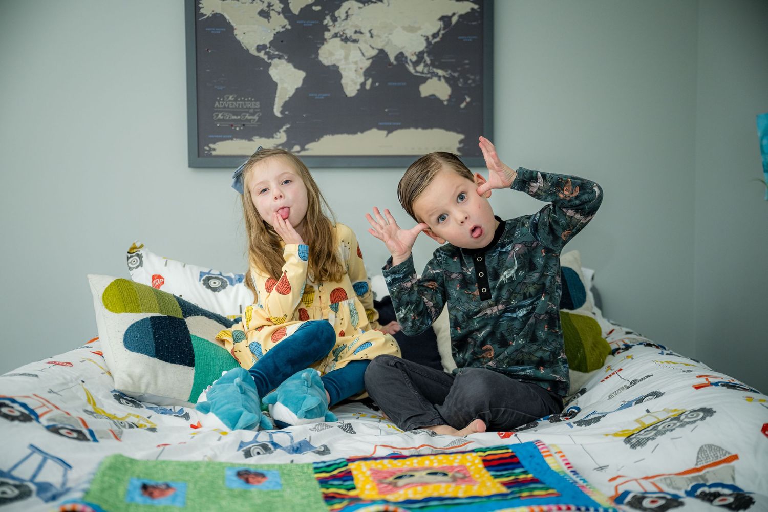 Two children sit on a bed with books and toys while posing playfully in front of a world map on the wall.