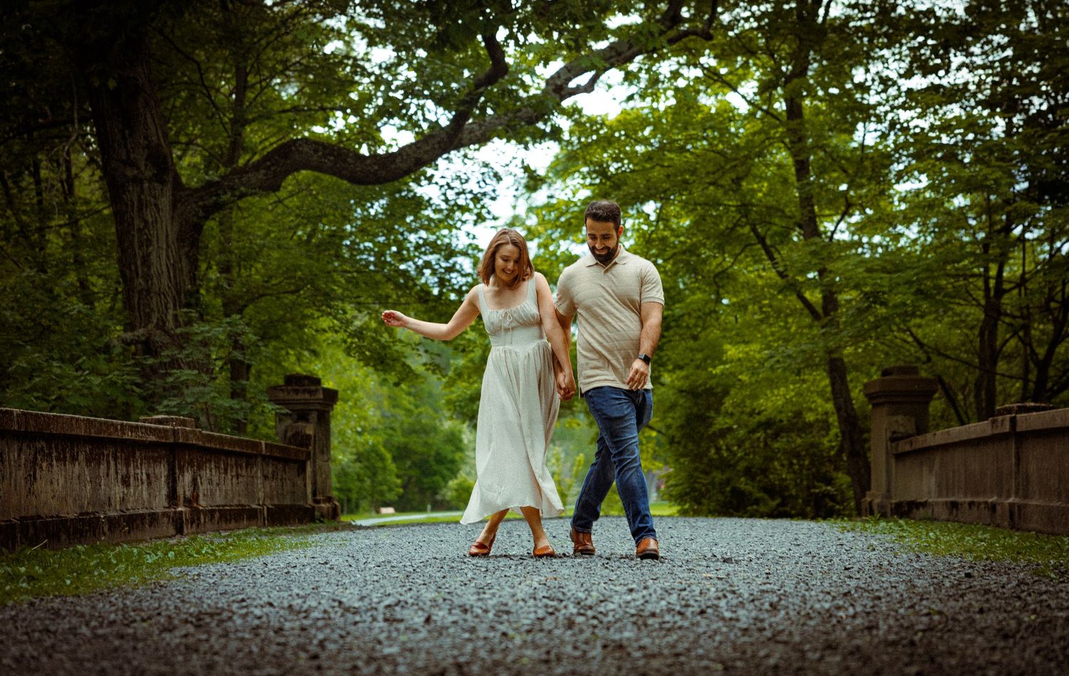 A couple stands together on a gravel path surrounded by lush green trees and an old stone bridge in a romantic setting.
