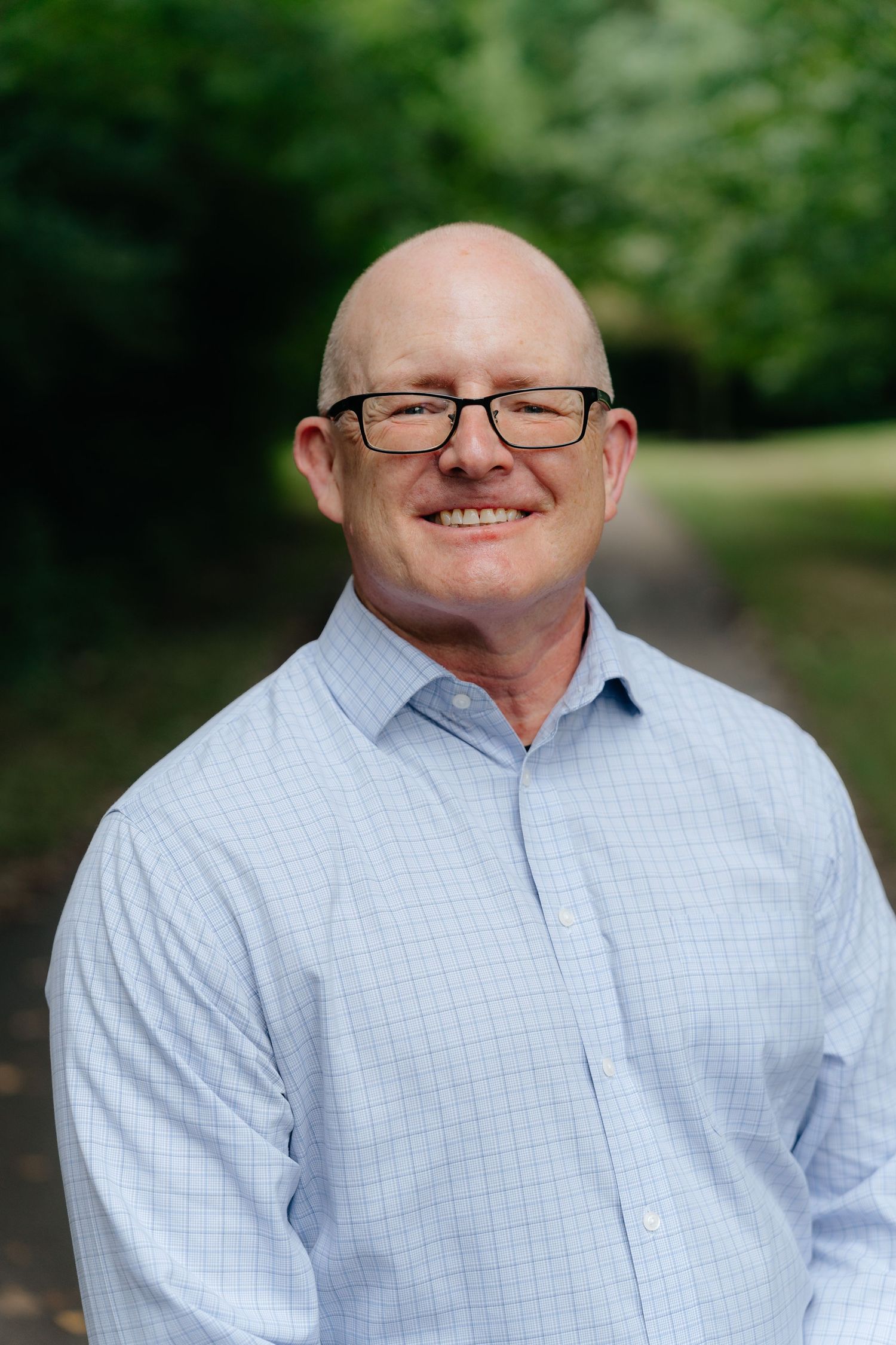 Professional headshot of businessman in light blue checkered shirt and glasses outdoors.