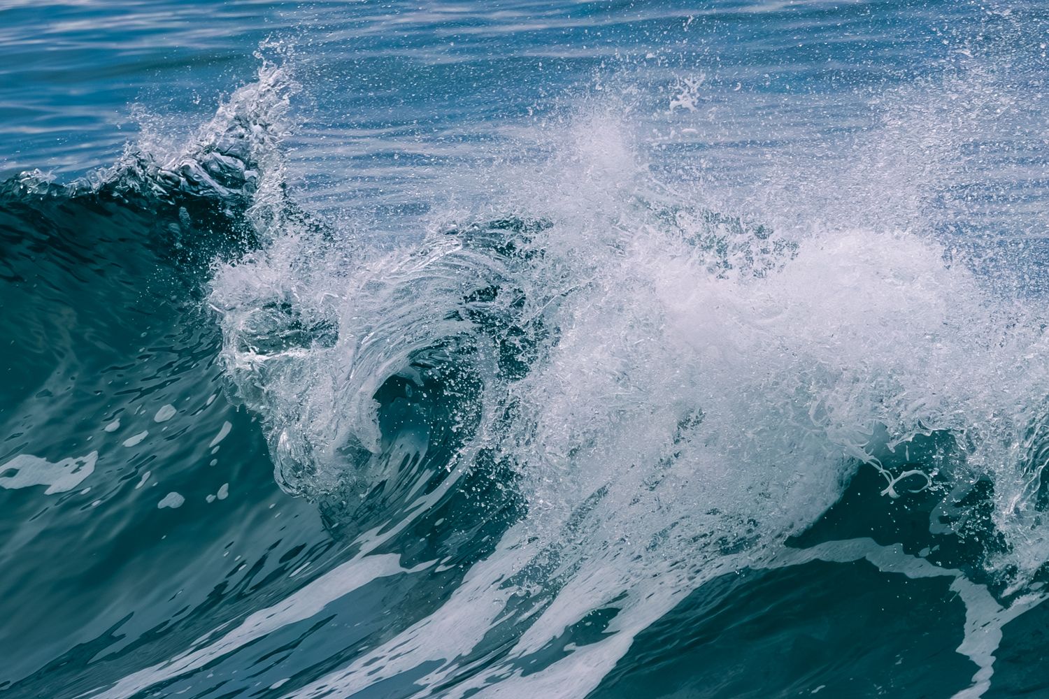 A powerful turquoise ocean wave crashes and sprays white foam against the deep blue water surface on Haida Gwaii.